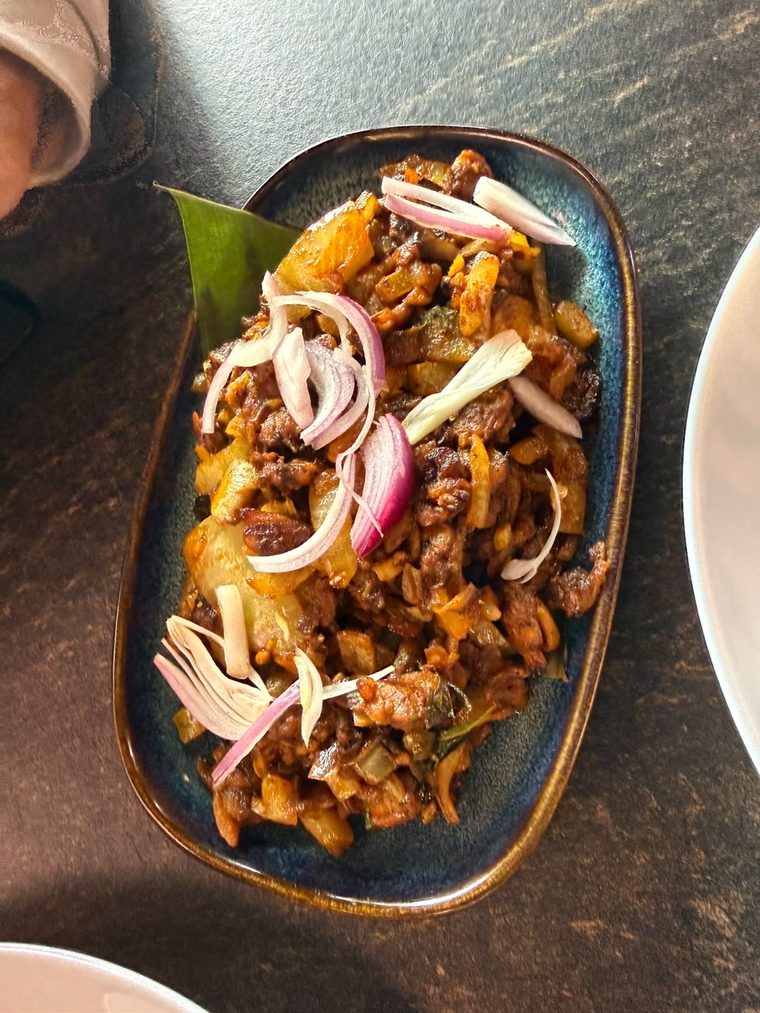 A dark stir-fried meat and coconut dish topped with raw onion rings, served over a banana leaf